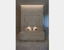 Luxuriöses Badezimmer mit freistehender Badewanne aus poliertem Naturstein mit auffälliger Maserung. Im Hintergrund ist eine Wandnische mit integrierter Beleuchtung und dekorativen Elementen zu sehen, daneben moderne Armaturen und helle Bodenfliesen.