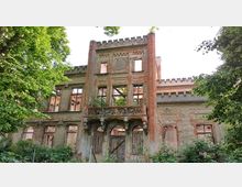 Fassade eines verfallenen historischen Gebäudes mit reich verziertem Balkon, freiliegendem Mauerwerk und spärlich bewachsenen Fensteröffnungen, umgeben von dichtem Grün und Bäumen. Das Gebäude zeigt Spuren von Verwitterung und einstigen architektonischen Details wie Zinnen und Ornamenten.