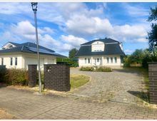 Einfahrt zu einem Wohnhaus mit heller Fassade und dunkel gedecktem Mansarddach, davor ein gepflasterter Hof und ein niedriges Blumenbeet. Links steht ein separates Garagengebäude mit Solarpaneelen auf dem Dach, am Straßenrand sind dunkle Ziegelpfeiler und eine Laterne zu sehen.