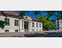 Historisches Gebäude mit der Aufschrift "Alte Forstakademie" über dem Eingang, flankiert von zwei Säulen und einem Dreiecksgiebel mit Verzierungen. Im Vordergrund ein gepflasterter Gehweg und Grünflächen, umgeben von Bäumen und benachbarten Wohnhäusern.