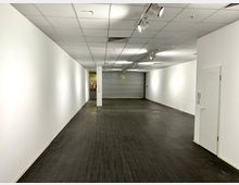 Leerer Innenraum mit weißen Wänden, dunklem Boden und einer abgehängten Decke mit Einbauleuchten. Im Hintergrund befindet sich ein geschlossenes Rolltor sowie eine Notausgangstür mit grünem Schild.