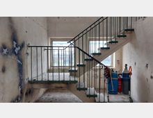 Treppe in einem verlassenen Gebäude mit Roststellen und abgeblättertem Putz. Im Hintergrund befinden sich zwei Fenster, durch die Tageslicht einfällt, und rechts unten steht eine Gasflaschenausrüstung auf einem Rollwagen.