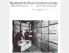 Schwarz-Weiß-Fotografie eines Kühlhauses aus dem frühen 20. Jahrhundert in Berlin mit beschrifteten Holzkisten, die in hohen Stapeln gelagert sind. Drei Männer in Arbeitskleidung inspizieren und verpacken gefrorenen Fisch in einer offenen Holzkiste.