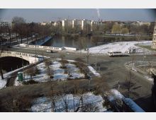 Straßenkreuzung in einer urbanen Winterlandschaft mit leichtem Schneebelag, einer Brücke über einen Fluss und angrenzenden Parkflächen mit kahlen Bäumen; im Hintergrund Wohngebäude, Baustellenkräne sowie der Fernsehturm erkennbar.
