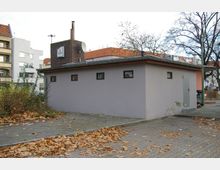 Flaches, rechteckiges Gebäude mit grauer Fassade und kleinen Fenstern, umgeben von einem gepflasterten Gehweg und herbstlichem Laub. Im Hintergrund sind mehrstöckige Wohnhäuser mit roten Dächern sowie Bäume zu sehen.