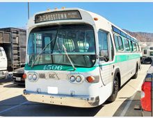 Ein klassischer weißer und türkisfarbener Linienbus mit der Beschriftung „3 - Crosstown Westwood Pico“ an der Front, geparkt auf einem sonnigen Parkplatz. Im Hintergrund sind mehrere Fahrzeuge und Hügel zu sehen.