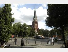 Straßenszene mit einer roten Backsteinkirche und hohem Spitzdach im Hintergrund, umgeben von Bäumen. Im Vordergrund befindet sich ein gepflasterter Platz mit Laternen, parkenden Fahrzeugen und Passanten.