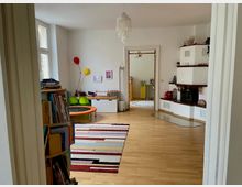 Wohnzimmer mit Holzboden, buntem Teppich und einer Spielecke mit kleinem Trampolin. Im Hintergrund ein gemauerter Kamin mit Holzregalen und eine offene Tür, die in einen angrenzenden Raum führt. Links ein Bücherregal und große Fenster, die Tageslicht hereinlassen.