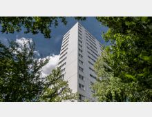 Hohes Wohngebäude mit weißer Fassade, umgeben von dichtem Grün und Bäumen, die den unteren Bereich des Bildes einrahmen; im Hintergrund ein bewölkter blauer Himmel.