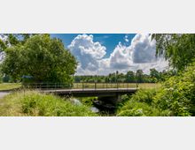 Brücke über einen kleinen, von grüner Vegetation umgebenen Kanal in einer ländlichen Landschaft mit Bäumen und Wiesen im Hintergrund, unter einem Himmel mit weißen Wolken. Ein Boot liegt unter der Brücke am Ufer des Kanals.