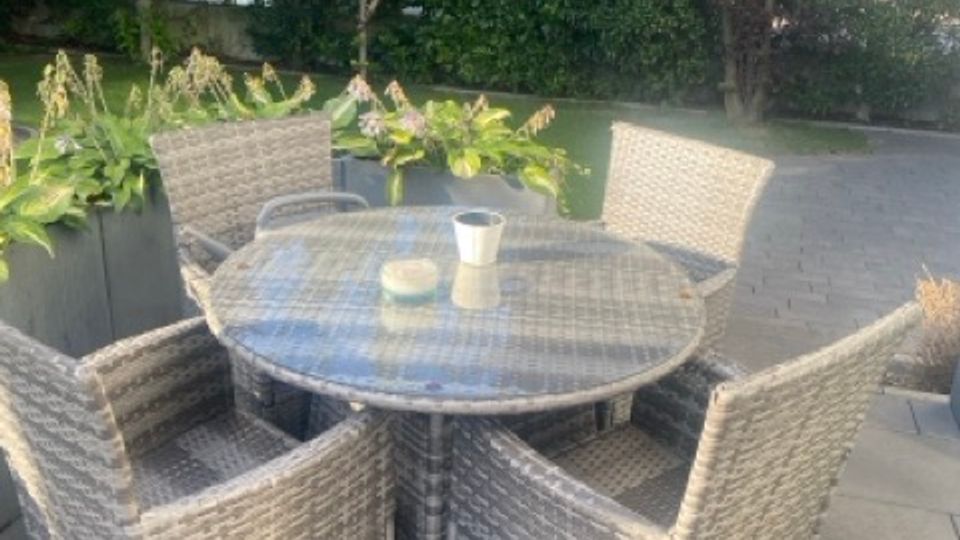 Runde Gartentischgruppe aus grauem Polyrattan mit vier Sesseln auf einer gepflasterten Terrasse. Im Hintergrund stehen große Pflanzkübel mit grünen Pflanzen sowie eine dichte Hecke und Bäume.