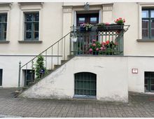 Fassade eines hellen, historischen Gebäudes mit grün gerahmten Fenstern. Eine Metalltreppe mit Geländer führt zu einem kleinen Balkon, der dicht mit bunten Blumen in Töpfen bepflanzt ist; darunter befindet sich ein vergittertes Kellerfenster.