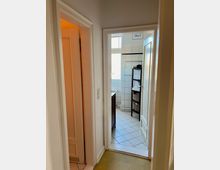 Blick in einen schmalen Flur, der zu einem hellen Badezimmer mit weißen Fliesen und einem Fenster führt. Rechts steht ein Regal mit Handtüchern und Körben, links führt eine weitere Tür in einen Raum mit warmer Beleuchtung.