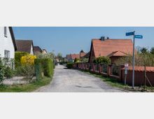 Wohnstraße in einem ruhigen Vorort mit Einfamilienhäusern, die rote Ziegeldächer und Gärten mit Hecken und Sträuchern haben. Ein unbefestigter Weg führt zwischen den Häuserreihen entlang, und Straßenschilder markieren die Kreuzung von "Milanweg" und "Hasenweg".