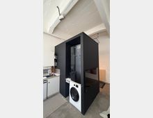 Innenansicht einer modernen Küche in einem Loft mit weiß gestrichenen Deckenbalken und Betonboden. Ein großer, schwarzer Einbaukubus integriert Kühlschrank und Waschmaschine; daneben stehen weiße Küchenschränke mit Spüle und Arbeitsfläche.