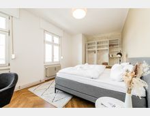 Helles Schlafzimmer mit großem grauem Polsterbett, weißen Wänden und hohen Fenstern mit Heizkörper darunter. Im Hintergrund stehen offene Regale vor einer roh verputzten Wand; Holzboden im Fischgrätmuster, kleiner Teppich und Nachttischlampe ergänzen die Einrichtung.