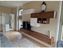 Moderne Wohnzimmergestaltung mit hellem Holzboden, einer beige gestrichenen Wand und hängenden Regalelementen aus Holz und Weiß. Ein Fernseher steht auf dem Sideboard, dekoriert mit einer Buddha-Büste und Orchideen, während ein großer Spiegel und Stauraummöbel den Raum ergänzen.