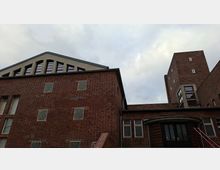 Backstein-Gebäude mit schlichtem, funktionalem Design, bestehend aus mehreren Segmenten mit kleinen Fenstern. Ein gewölbter Eingangsbogen sowie ein hoher Turm im Hintergrund dominieren die Architektur.