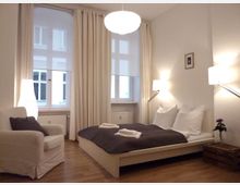 Helles Schlafzimmer mit einem Doppelbett, weißen Bettwäschen und dunkler Decke, einer weißen Sitzgelegenheit, Holzfußboden und großen Fenstern mit Vorhängen. Der Raum ist mit Nachttischlampen und einer Stehlampe beleuchtet.