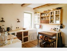 Gemütliche Landhausküche mit cremefarbenen Wänden, sichtbaren Holzbalken und einem großen Fenster, das Tageslicht hereinlässt. Die Ausstattung umfasst eine rustikale Arbeitsfläche mit Fliesen, offene Holzschränke mit Glasfronten, Kupfertöpfen auf einem mobilen Küchenwagen und eine Spüle unter dem Fenster.