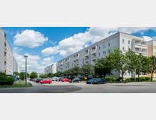 Mehrstöckige Wohngebäude aus Beton mit Balkonen säumen eine breite Straße, entlang der auf beiden Seiten Autos geparkt sind. Bäume und Grünflächen befinden sich vor den Gebäuden, unter einem Himmel mit weißen Wolken.