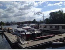 Ein kleiner Hafenbereich mit mehreren festgemachten Booten und Hausbooten an Holzstegen, umgeben von Bäumen und Gebäuden im Hintergrund. Ein Schornstein, der Rauch ausstößt, ist deutlich sichtbar vor einem bewölkten Himmel.