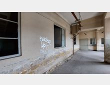 Verlassener Innenraum mit beschädigten Wänden, auf denen Graffiti sichtbar sind. Die Decke enthält alte Rohrleitungen und Lampen, während Fenster und eine geschlossene Tür den Raum zusätzlich kennzeichnen.