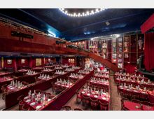 Ein elegantes Theaterrestaurant mit in Reihen angeordneten, festlich gedeckten Tischen, roten Samtpolstern und einer Bühne auf der rechten Seite. Der Raum verfügt über eine Wand mit ausgestellten Kostümen und Plakaten sowie einen oberen Balkonbereich mit Beleuchtungs- und Technikgeräten.