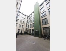 Innenhof eines mehrstöckigen Gebäudes mit weißen Ziegelwänden und großen Fenstern. Ein vertikaler, grüner Anbau mit Tür dominiert die rechte Seite, während Fahrräder entlang der Wände abgestellt sind.