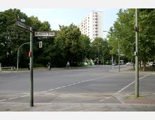 Straßenkreuzung mit Straßenschildern "Paulsborner Straße" und "Westfälische Straße", umgeben von Bäumen und einem hohen Wohngebäude im Hintergrund. Mehrere Verkehrsteilnehmer, darunter Radfahrer und Autos, sind auf der Straße zu sehen.