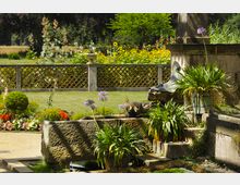 Ein gepflegter Garten mit einem steinernen Brunnen, verziert durch eine Fischfigur, umgeben von bunten Blumenbeeten und grünen Pflanzen. Im Hintergrund steht eine schmuckvolle Steinbrüstung mit rautenförmigen Mustern und weitere Blumenrabatten vor einer Wiesenlandschaft.