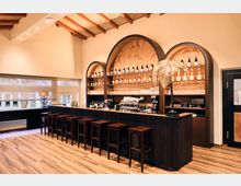 A modern bar interior with a wooden counter lined with high stools and an arched shelving unit displaying bottles and glassware. The warm space features a patterned backdrop, wooden ceiling beams, and a decorative golden fern centerpiece.