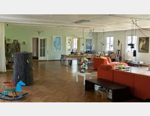 Großer, heller Raum mit Holzparkettboden, genutzt als Wohn- und Arbeitsbereich. Der Raum enthält eine rote Couch, mehrere Tische mit Stühlen, Regale, Kunstwerke an den Wänden und dekorative Gegenstände wie ein buntes Schaukelpferd und eine bemalte Skulptur; mehrere Fenster lassen Tageslicht herein.