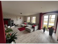 Wohnzimmer mit moderner Einrichtung, einem grauen Ecksofa, einem Kamin an der linken Wand und großen Fenstern mit Blick nach draußen. Der Raum ist hell gestaltet, mit roten Akzentwänden, einem Holzboden und mehreren Zimmerpflanzen.