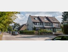 Straßenszene in einem Wohngebiet mit freistehenden und Reihenhäusern aus hellem Backstein, schrägen Dächern und Dachfenstern. Im Vordergrund geparkte Autos entlang der Straße, umgeben von Bäumen und gepflegten Vorgärten.