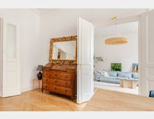 Heller Wohnungsflur mit Fischgrätparkett und weißen Flügeltüren, neben einer antiken Holzkommode mit großem, goldverziertem Spiegel. Durch die geöffnete Tür ist ein Wohnzimmer mit hellblauem Sofa, Glastisch und runder Deckenleuchte zu sehen.