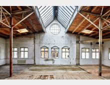 Alter, offener Innenraum eines Industriegebäudes mit Holzdielenboden, unverputzten weißen Backsteinwänden und sichtbarer Holzkonstruktion an der Decke. Große Fenster und ein rundes Oberlicht sorgen für natürliches Licht.