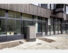 Moderne Gewerbe- oder Bürogebäude mit einer Fassade aus großen Glasfenstern und dunklem Holzverkleidungen. Im Vordergrund ist ein gepflasterter Bereich mit einem Lüftungsschacht sowie schmalen, unbepflanzten Beeten sichtbar.