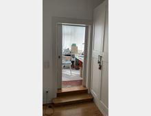 Blick durch eine Türöffnung in ein helles Arbeitszimmer mit weißem Schreibtisch, weißem Stuhl und mehreren kleinen Regalen. Holzboden im Vordergrund, niedrige Stufe zum Arbeitszimmer mit rotbraunen Fliesen und einem gestreiften Teppich.