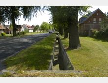 Straßenszene in einer ländlichen Umgebung mit einer asphaltierten Fahrbahn, gesäumt von Bäumen und niedrigeren Wohnhäusern aus Backstein und Putz. Im Vordergrund verläuft ein offener Entwässerungsgraben aus Beton entlang der Wiese.