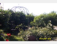 Ein Garten mit blühenden Sträuchern und einem Pavillon aus schwarzem Metall mit geschwungenen Verzierungen, umgeben von dichtem Laubwerk. Im Vordergrund befinden sich weiße und rote Rosen sowie ein großer Stein.