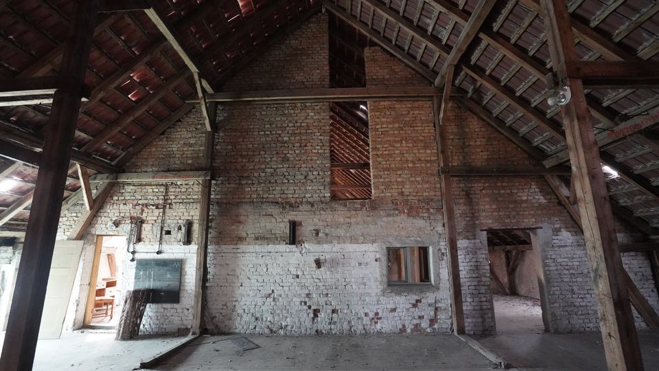 Innenraum einer alten Scheune mit hohen Holzbindern und sichtbarer Dachkonstruktion über einer teils weiß gestrichenen, teils unverputzten Backsteinwand. Mehrere Öffnungen und Türen führen in Nebenräume, der Boden besteht aus rohem Beton mit etwas Schmutz und Geröll.