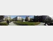 Ein Park mit einem zentralen Brunnen, umgeben von Blumenbeeten und Rasenflächen, eingefasst von einem Gehweg. Im Hintergrund stehen mehrstöckige Wohn- und Geschäftsgebäude sowie vereinzelte Bäume.