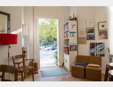 Ein gemütlicher Wohnraum mit Holzboden, dekoriert mit Stühlen, Regalen voller Bücher und Bildern an den Wänden. Die offene Tür führt zu einer Straße mit grünen Bäumen und geparkten Autos.