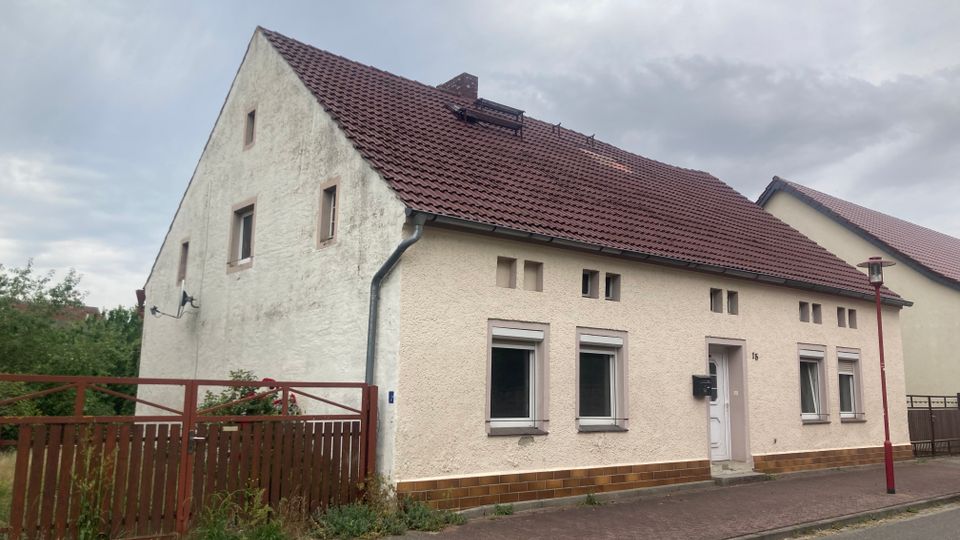 Ein einstöckiges Wohnhaus mit beigefarbener Fassade, roten Dachziegeln und mehreren rechteckigen Fenstern. Vor dem Haus befindet sich ein kleiner Zaun, und die Straße ist mit Gehwegplatten ausgelegt.