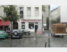 Straßenszene mit einer Fahrschule im Erdgeschoss eines Wohngebäudes; vor dem Gebäude sind Autos und ein Motorrad geparkt, daneben ein Lieferwagen mit der Aufschrift „Autovermietung Köhrich“. Ein Baum und eine Straßenlaterne stehen am Rand eines nassen Pflasters.