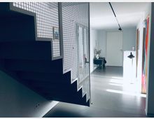 Moderner Innenraum mit einer freitragenden Treppe, die von einem Metallgitter eingefasst ist. Der Raum ist minimalistisch gestaltet mit glattem Betonboden, weißen Wänden und einzelnen Möbelstücken wie einem kleinen Beistelltisch und Dekorationen.