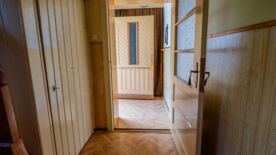 Schmaler Flur in einem älteren Wohnhaus mit Holzvertäfelung, Einbauschrank links und Parkett im Fischgrätmuster. Im Vordergrund steht eine halb geöffnete Tür mit strukturiertem Glas, die den Blick in einen angrenzenden Raum mit weiterer Innentür und Vorhang freigibt.