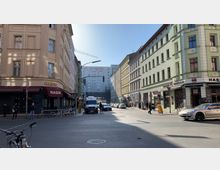 Straßenszene in einer urbanen Umgebung mit mehrstöckigen, historischen Gebäuden in Creme- und Grüntönen. Im Vordergrund ein Restaurant mit der Aufschrift „Hasir“, parkende Autos, Fußgänger und ein Lieferwagen; im Hintergrund ein modernes Gebäude mit der Aufschrift „ADHS“.