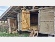 Holz- und Lehmbauernhof mit offenstehender hölzerner Tür, die zu einem Innenraum mit Stroh führt. Die Konstruktion besteht aus grob behauenen Holzstämmen, Lehmwänden und einem schrägen Dach; im Vordergrund liegt gestapeltes Holz und ein Zaun aus dünnen Ästen grenzt den Bereich ab.